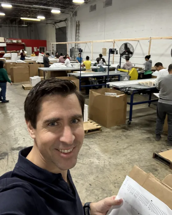Bernhard Brandl on a production floor beside assembly benches, holding a printed process sheet.